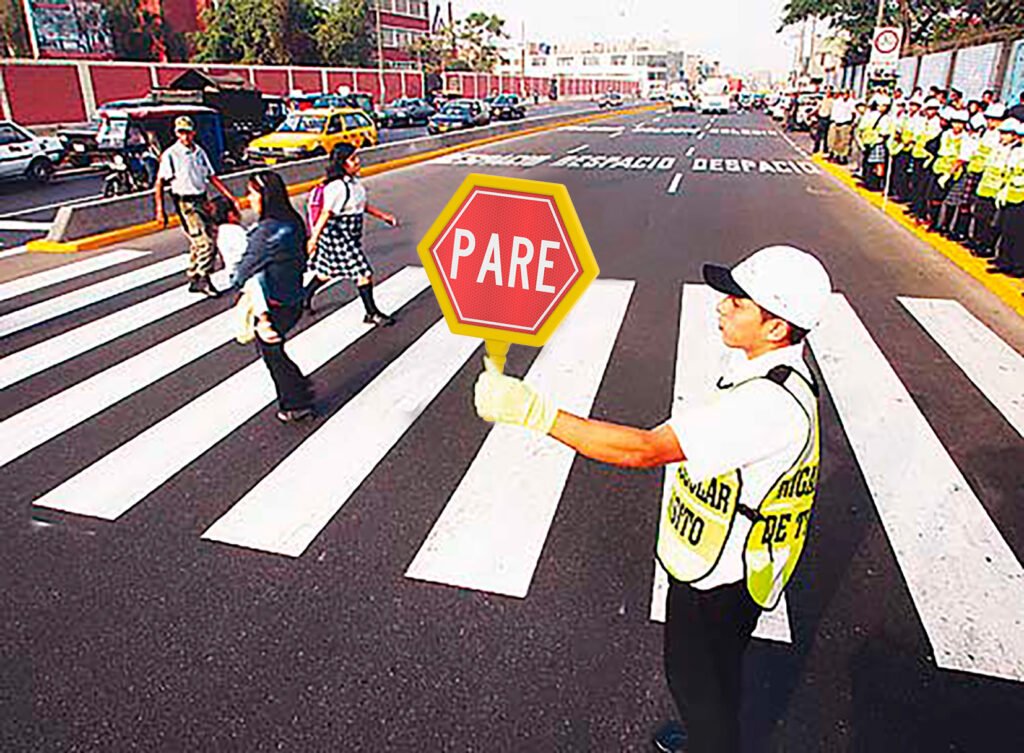 Agente de tránsito sostiene paleta de “PARE” para detener vehículos mientras peatones cruzan de forma segura por paso peatonal en una avenida concurrida, destacando el control vial y la protección del peatón en zonas escolares o de alto flujo.








