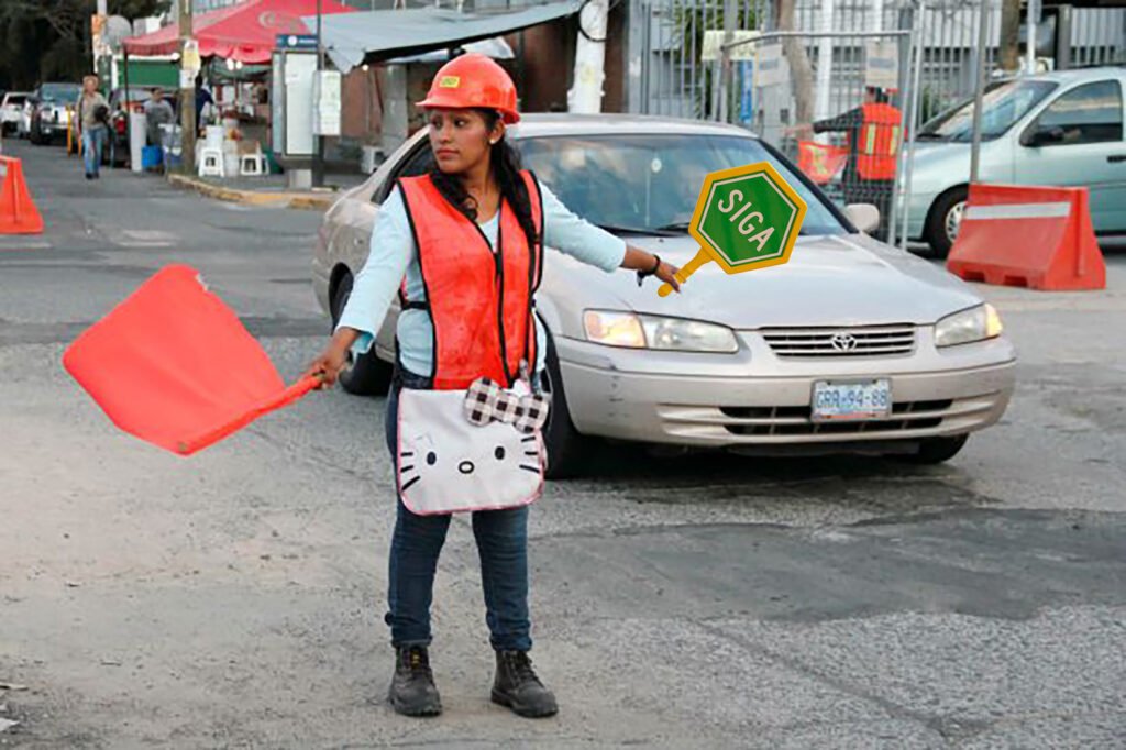 Trabajadora vial con casco y chaleco anaranjado dirige el tránsito en una zona de obra, utilizando una bandera roja y una paleta de “SIGA” para indicar el paso a los vehículos, promoviendo la seguridad y el control en áreas de mantenimiento urbano.